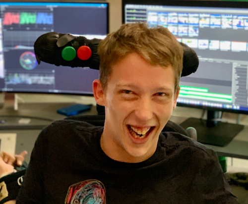 The young man smiles to camera from his wheelchair. A red switch and a green switch are mounted behind his head on his headrest. In the background is his edit suite, displaying video vector-scopes.